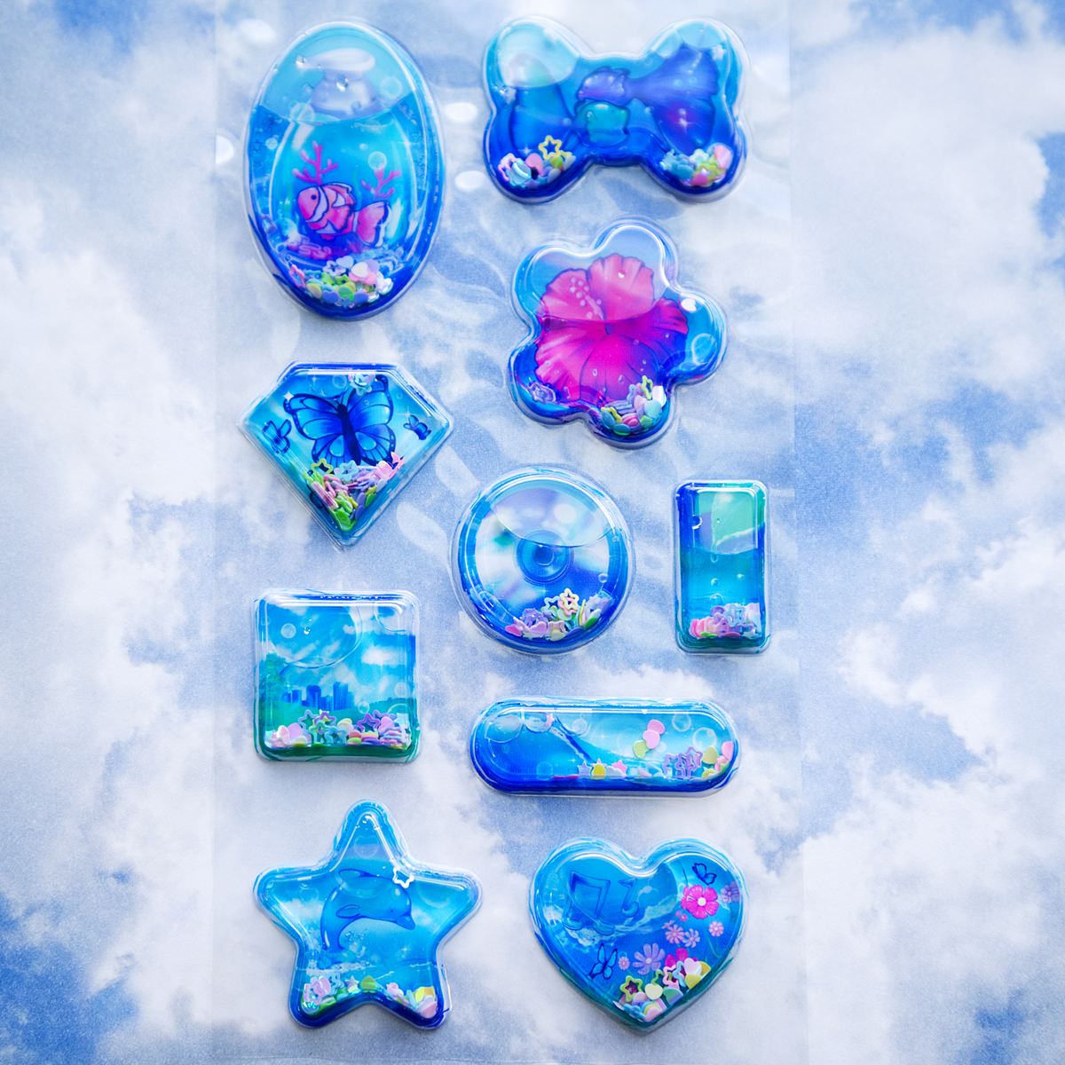 A sheet of clear plastic stickers filled with water and glitter confetti, featuring various Frutiger Aero themed illustrations including fish, butterflies, and flowers. The sheet is arranged on a transparent plastic layer, and is set on a sky-themed backdrop