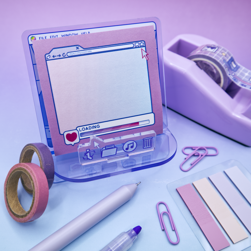 Pink sticky note pad with an internet browser loading screen design, sitting on an acrylic stand with a computer theme, on a purple background with stationery items.