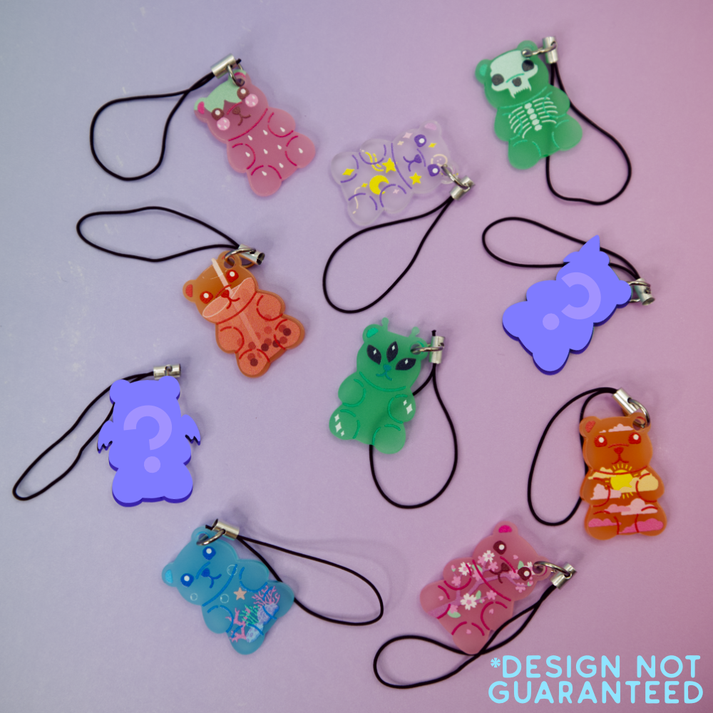 Colorful gummy bear-shaped phone charms on a light purple background with 'Design Not Guaranteed' text.  Two of the gummies are "mystery" designs and are silhouetted in purple with a question mark.