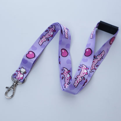 A pastel purple heart and "stay weird" text patterned lanyard with a breakaway safety buckle and silver lobster clasp sits on a white background. 