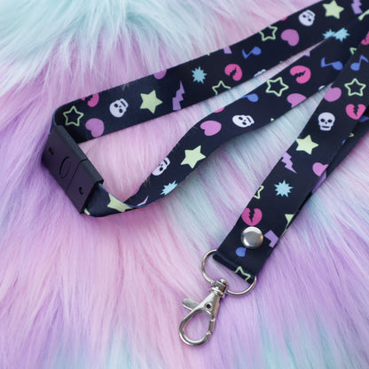 A black, early 2000's "scene kid" patterned lanyard with a breakaway safety buckle and silver lobster clasp sits on a fuzzy teal and pink background. 