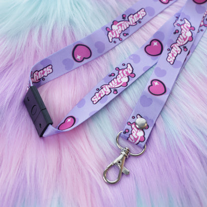 A pastel purple heart and "stay weird" text patterned lanyard with a breakaway safety buckle and silver lobster clasp sits on a fuzzy teal and pink background. 