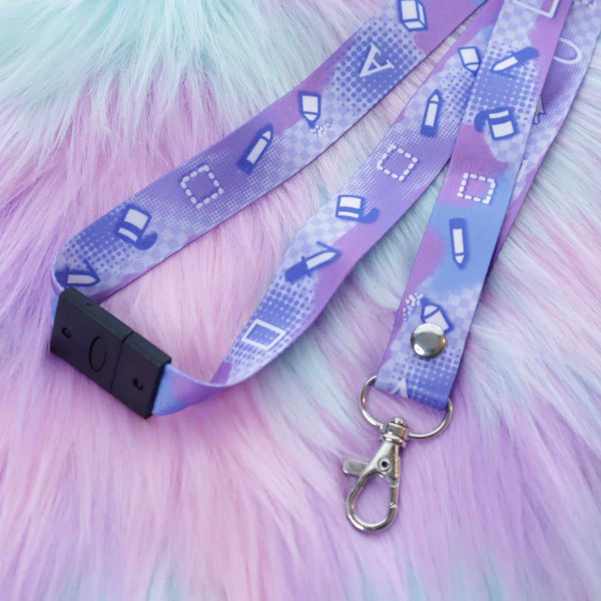A pastel purple digital art tool patterned lanyard with a breakaway safety buckle and silver lobster clasp sits on a fuzzy teal and pink background.  The safety buckle is opened to show the mechanism inside.