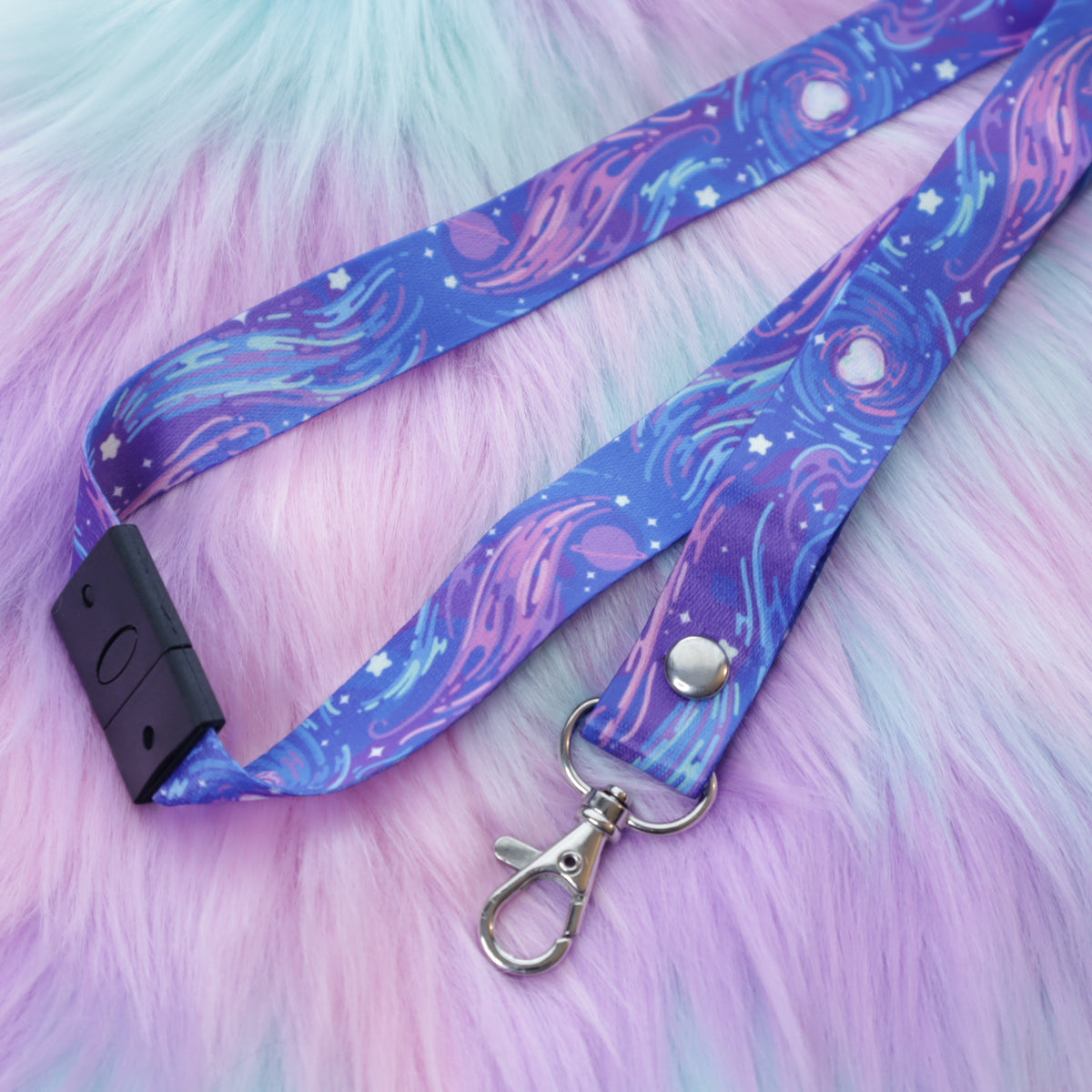 A purple space-patterned lanyard with a breakaway safety buckle and silver lobster clasp sits on a fuzzy teal and pink background.