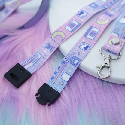 A pastel pink and blue computer UI patterned lanyard with a breakaway safety buckle and silver lobster clasp sits on a white tray above a fuzzy teal and pink background.  The safety buckle is opened to show the mechanism inside.