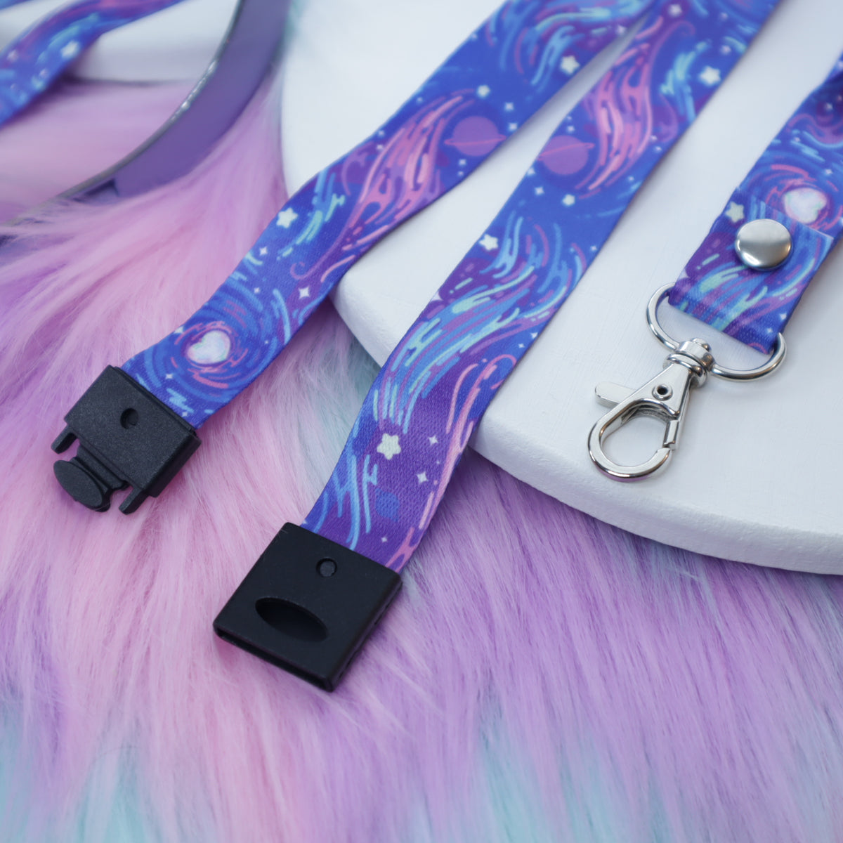 A purple space-patterned lanyard with a breakaway safety buckle and silver lobster clasp sits on a white tray above a fuzzy teal and pink background.  The safety buckle is opened to show the mechanism inside.