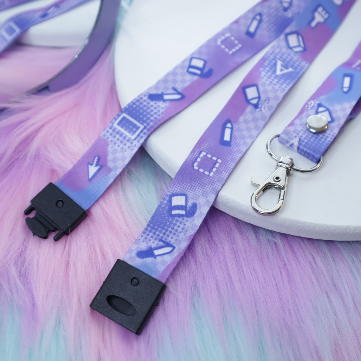 A pastel purple digital art tool patterned lanyard with a breakaway safety buckle and silver lobster clasp sits on a white tray above a fuzzy teal and pink background.  The safety buckle is opened to show the mechanism inside.