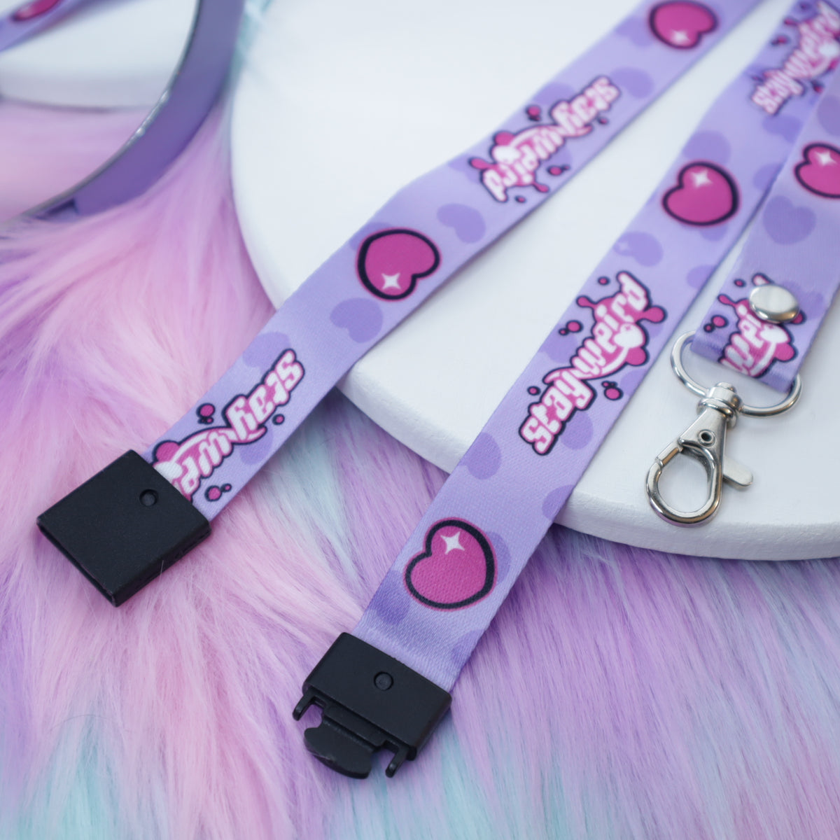 A pastel purple heart and "stay weird" text patterned lanyard with a breakaway safety buckle and silver lobster clasp sits on a white tray above a fuzzy teal and pink background.  The safety buckle is opened to show the mechanism inside.