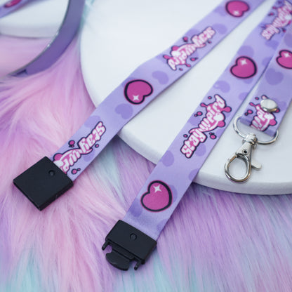 A pastel purple heart and "stay weird" text patterned lanyard with a breakaway safety buckle and silver lobster clasp sits on a white tray above a fuzzy teal and pink background.  The safety buckle is opened to show the mechanism inside.
