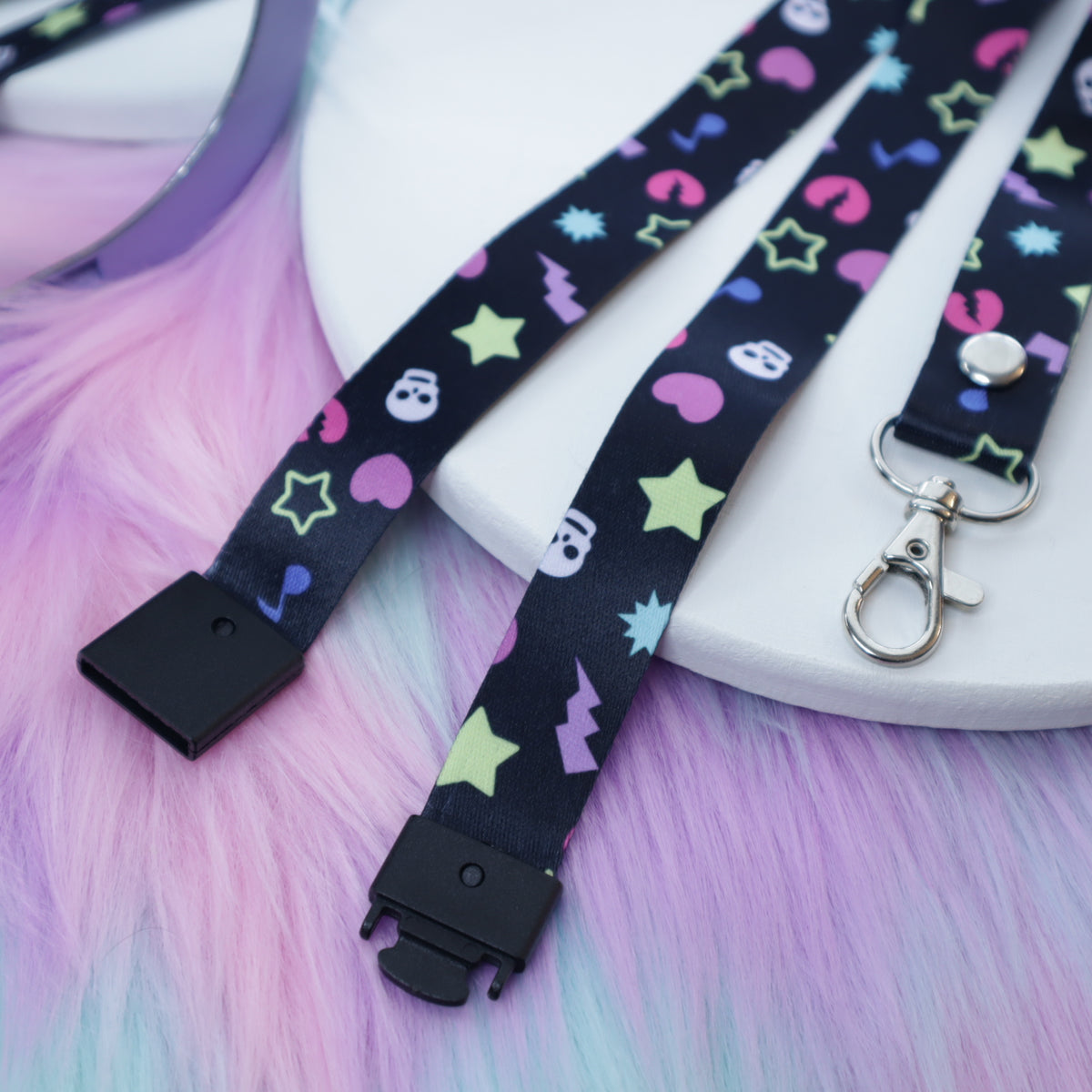 A black, early 2000's "scene kid" patterned lanyard with a breakaway safety buckle and silver lobster clasp sits on a white tray above a fuzzy teal and pink background.  The safety buckle is opened to show the mechanism inside.