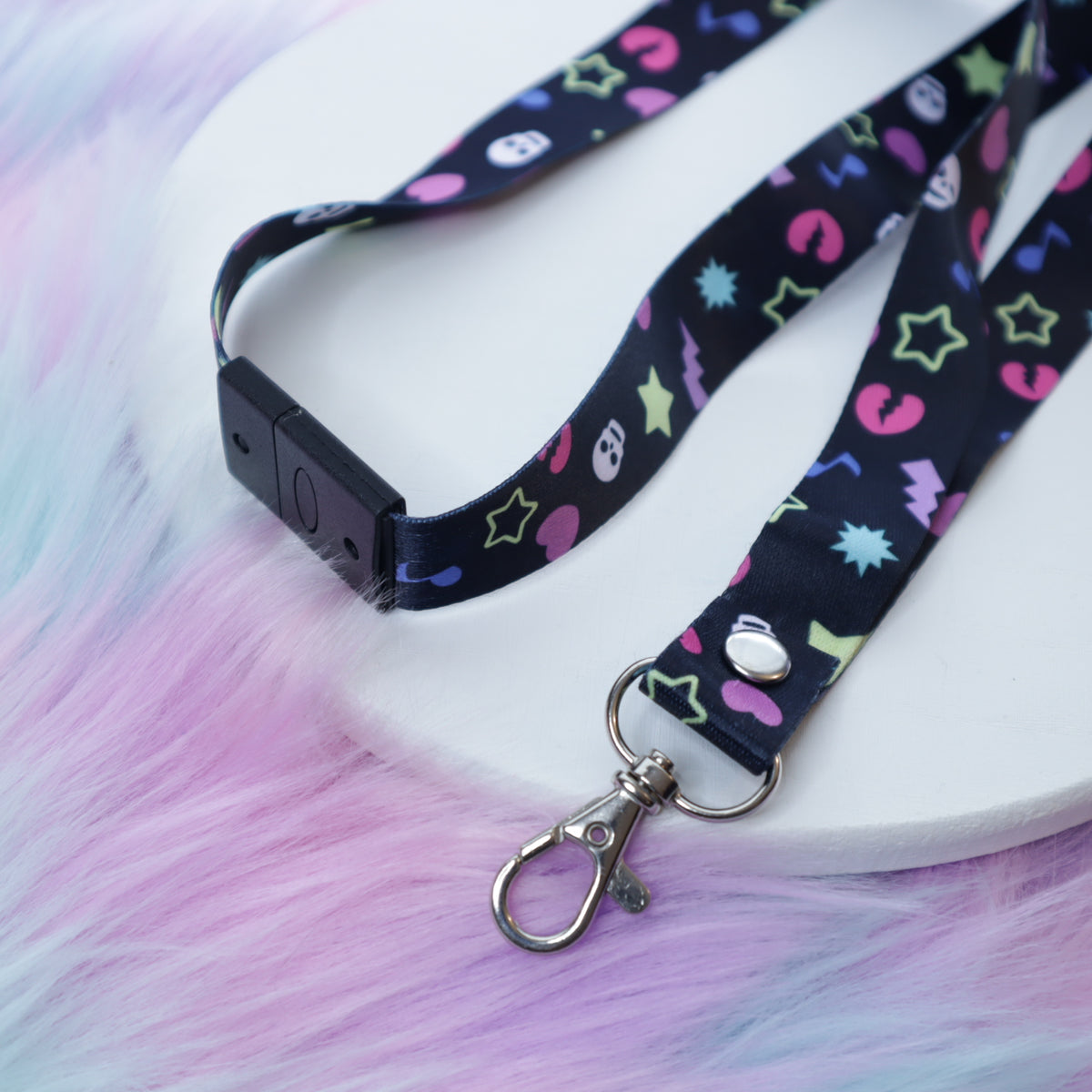 A black, early 2000's "scene kid" patterned lanyard with a breakaway safety buckle and silver lobster clasp sits on a white tray above a fuzzy teal and pink background. 