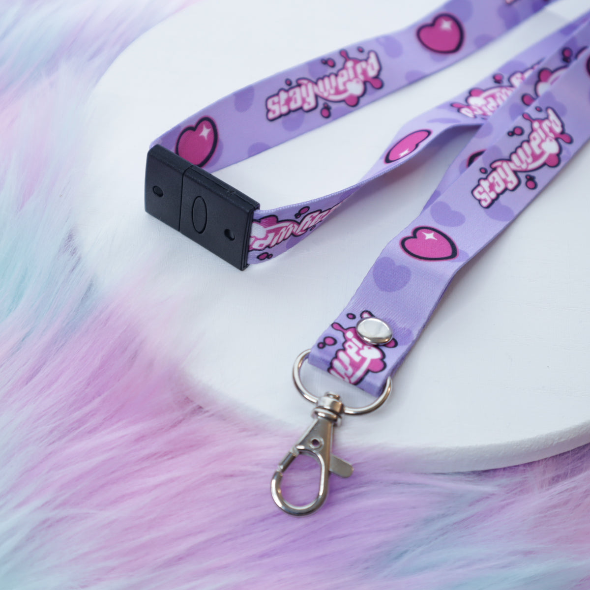 A pastel purple heart and "stay weird" text patterned lanyard with a breakaway safety buckle and silver lobster clasp sits on a white tray above a fuzzy teal and pink background. 