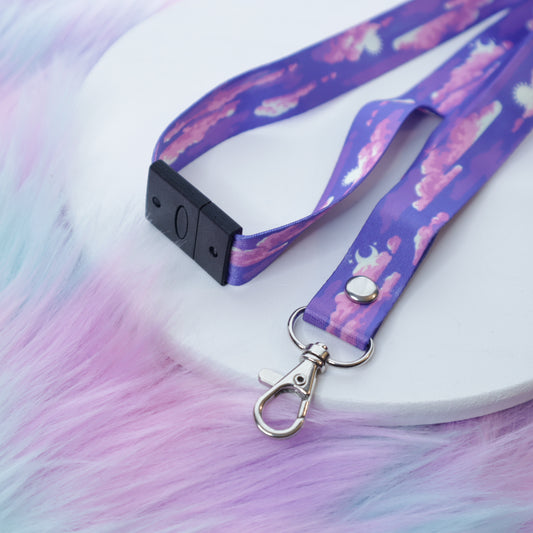 A purple cloud-patterned lanyard with a breakaway safety buckle and silver lobster clasp sits on a white tray above a fuzzy teal and pink background.
