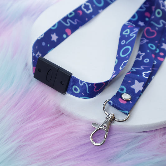 A blue arcade-carpet-patterned lanyard with a breakaway safety buckle and silver lobster clasp sits on a white tray above a fuzzy teal and pink background.