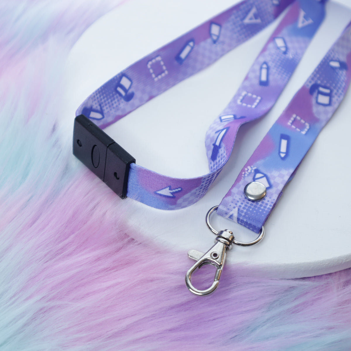 A pastel purple digital art tool patterned lanyard with a breakaway safety buckle and silver lobster clasp sits on a white tray above a fuzzy teal and pink background.
