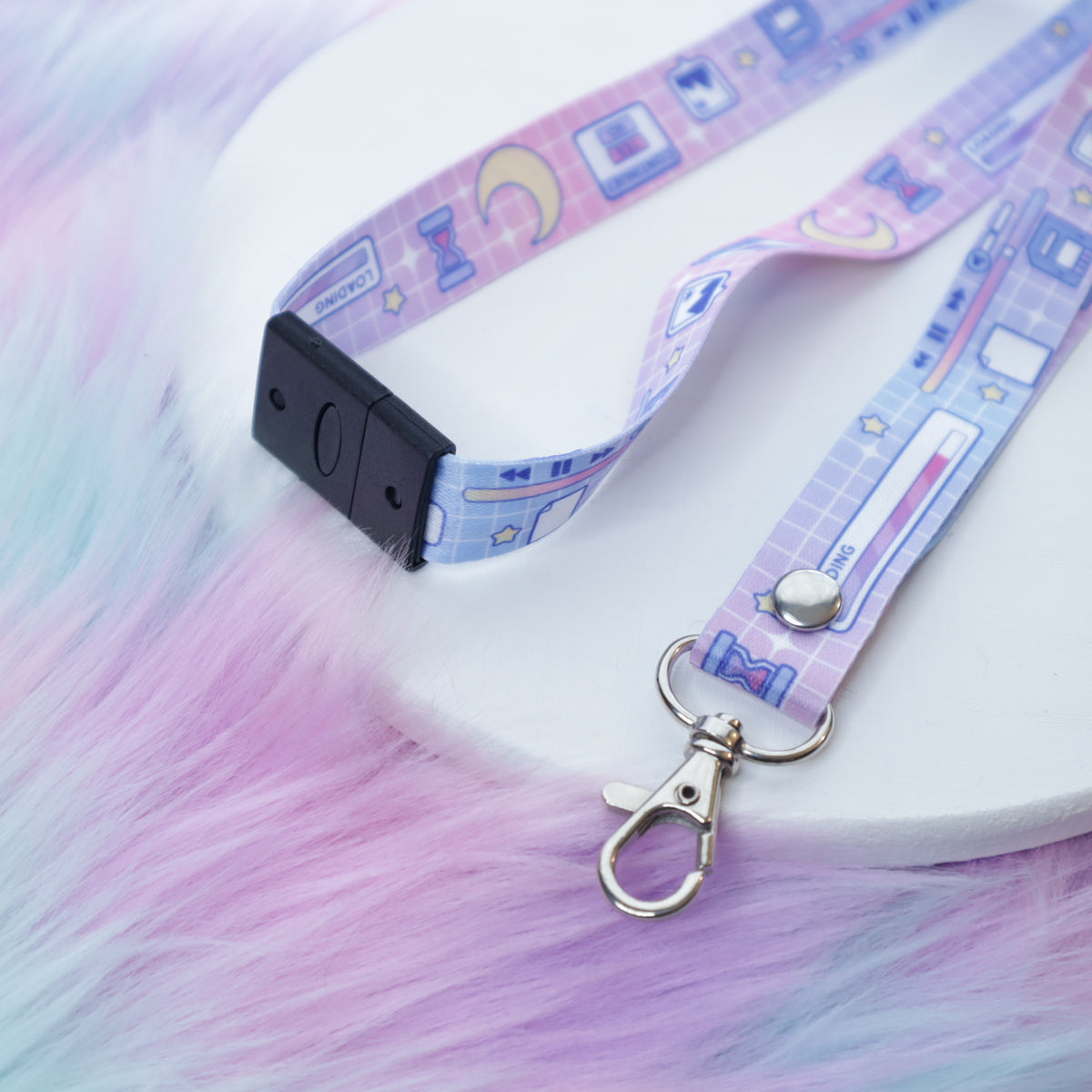 A pastel pink and blue computer UI patterned lanyard with a breakaway safety buckle and silver lobster clasp sits on a white tray above a fuzzy teal and pink background.