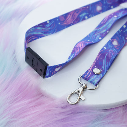 A purple space-patterned lanyard with a breakaway safety buckle and silver lobster clasp sits on a white tray above a fuzzy teal and pink background. 