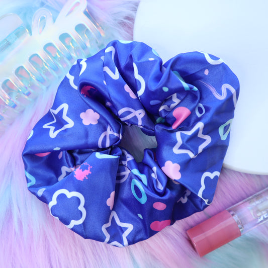 A blue "arcade pattern" satin scrunchie sits on a fuzzy pink background with a claw-clip hair accessory and lip gloss nearby