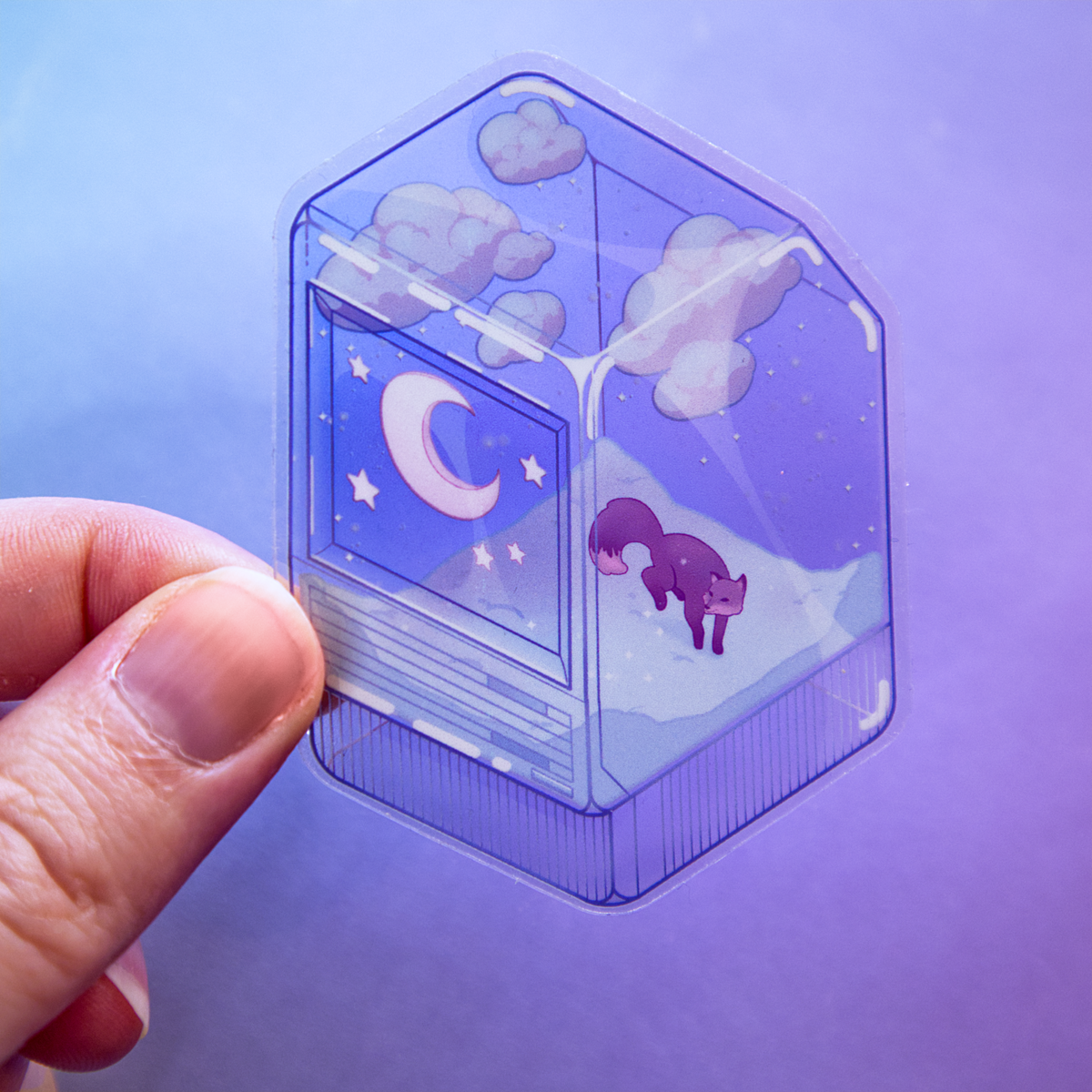 A hand holds a die-cut sticker featuring a winter scene with a small fox, night sky, and snow, inside of a translucent PC tower like a snow globe.  Sticker is photographed on a purple background.