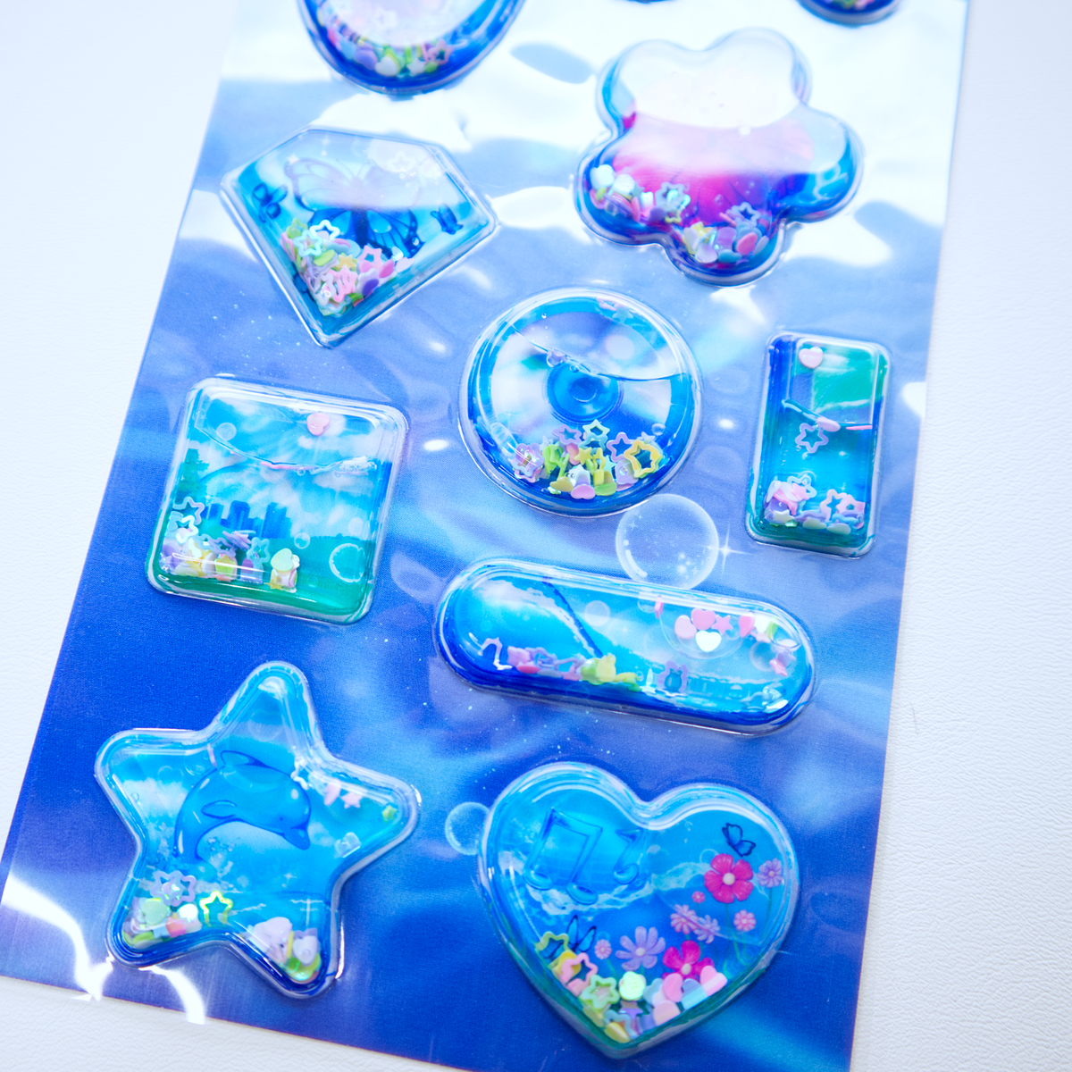 A sheet of clear plastic stickers filled with water and glitter confetti, featuring various Frutiger Aero themed illustrations including dolphins, butterflies, and flowers. 