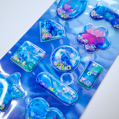 A sheet of clear plastic stickers filled with water and glitter confetti, featuring various Frutiger Aero themed illustrations including fish, butterflies, and flowers.