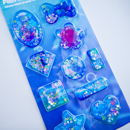 A sheet of clear plastic stickers filled with water and glitter confetti, featuring various Frutiger Aero themed illustrations including fish, butterflies, and flowers. 