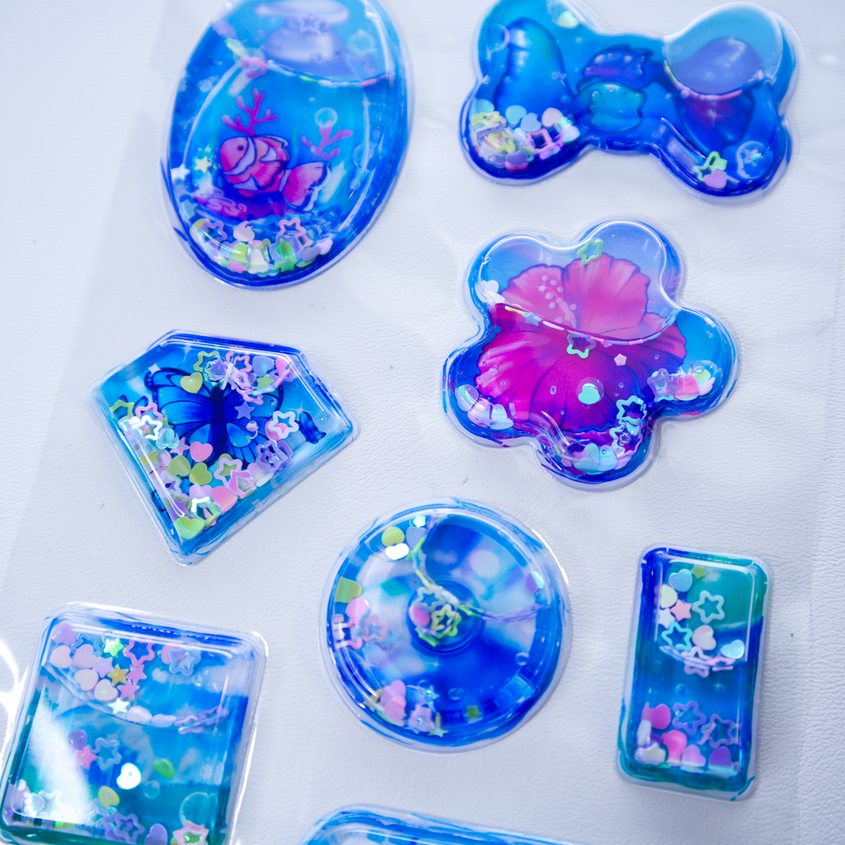 A sheet of clear plastic stickers filled with water and glitter confetti, featuring various Frutiger Aero themed illustrations including fish, butterflies, and flowers.
