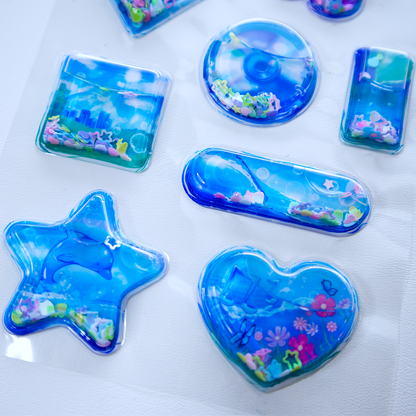 Close up of a sheet of clear plastic stickers filled with water and glitter confetti, featuring various Frutiger Aero themed illustrations including fish, butterflies, and flowers