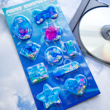 A sheet of clear plastic stickers filled with water and glitter confetti, featuring various Frutiger Aero themed illustrations including fish, butterflies, and flowers. A blank CD is shown on the right as a size reference.