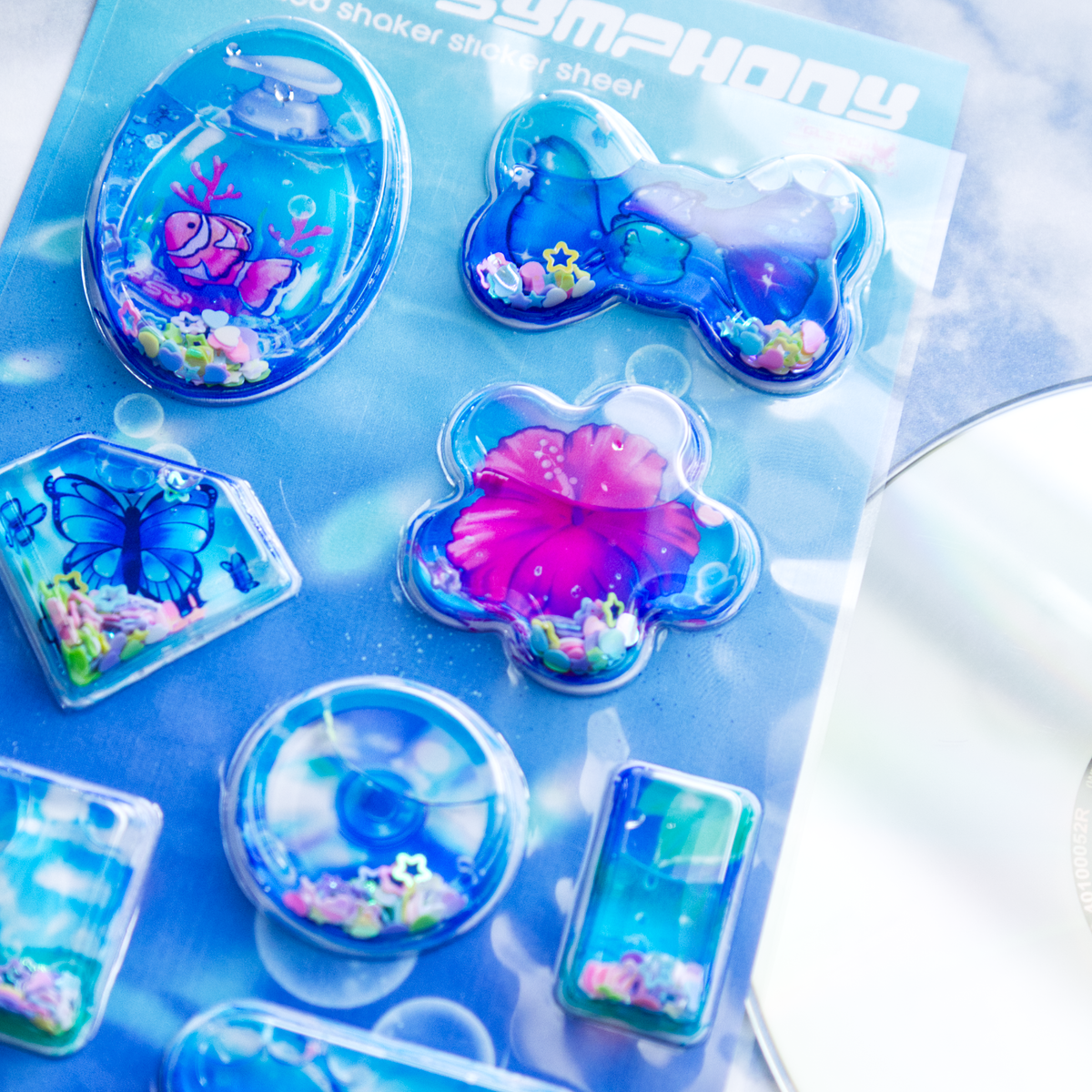 Close up of a sheet of clear plastic stickers filled with water and glitter confetti, featuring various Frutiger Aero themed illustrations including fish, butterflies, and flowers.