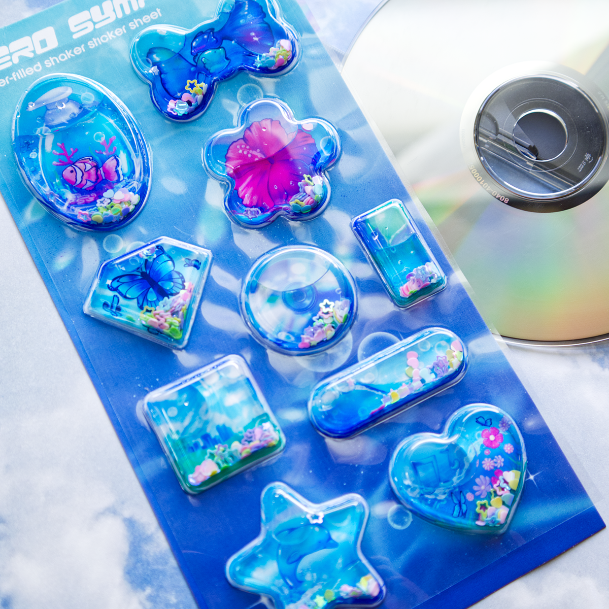 A sheet of clear plastic stickers filled with water and glitter confetti, featuring various Frutiger Aero themed illustrations including fish, butterflies, and flowers. A blank CD is shown on the right as a size reference.